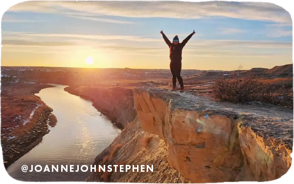 Woman standing on a cliff with her arms outstretched; a river is below and sunset is in the background, with overlay text that reads: @JOANNEJOHNSTEPHEN