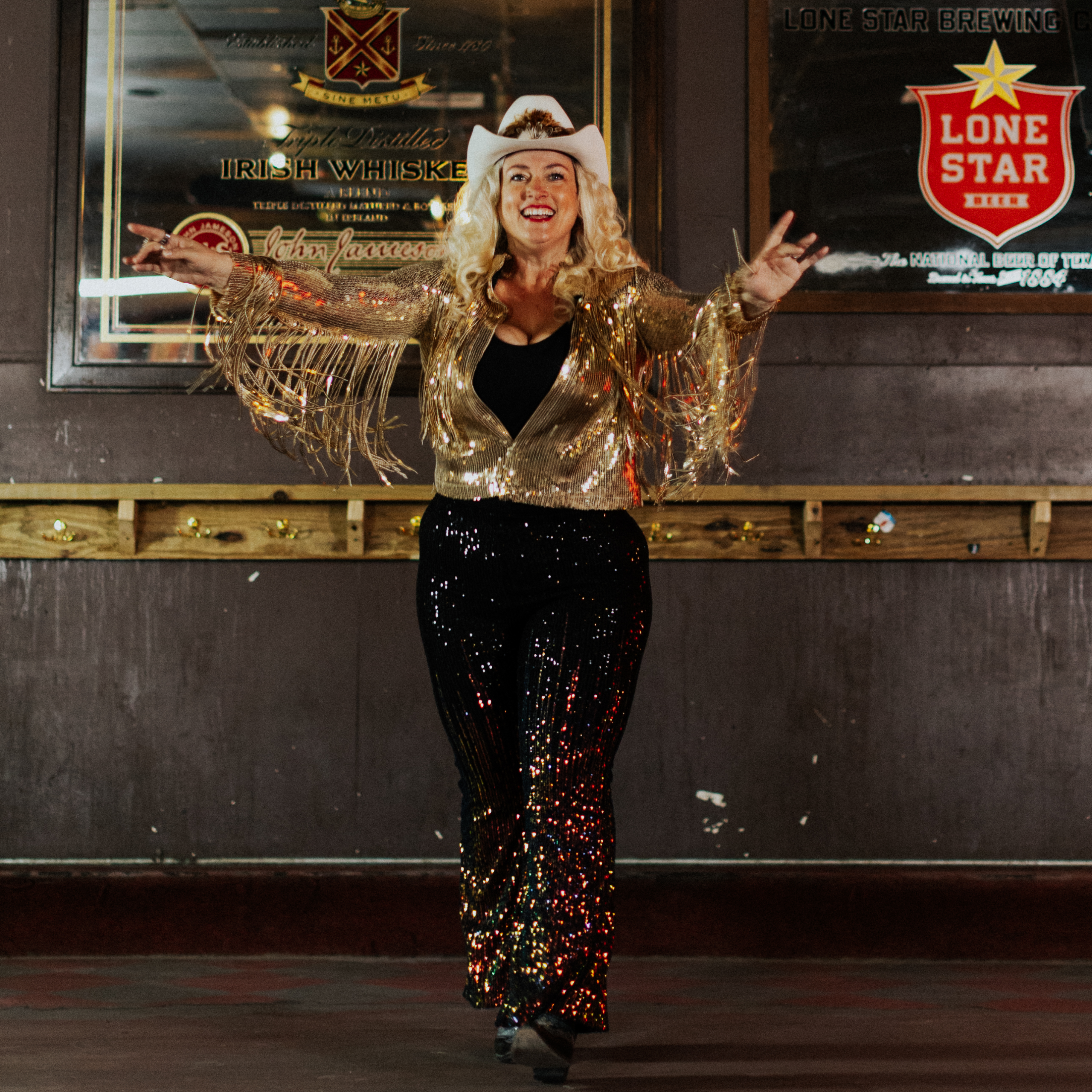 Woman wearing a cowboy hat and boots, sparkling gold fringe jacket and dark sequined pants strikes a dance pose 