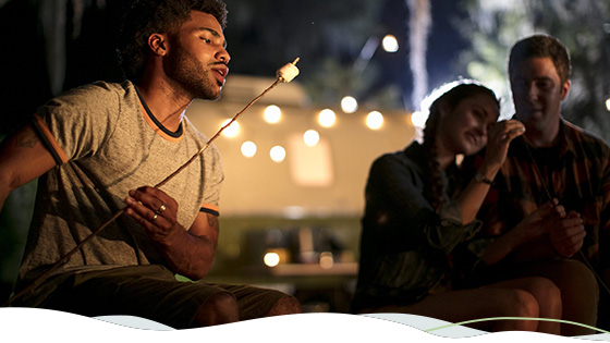 A man roasts a marshmallow, with twinkling lights and a camper in the background.