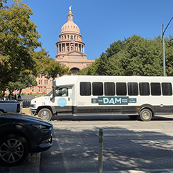 The white Dam Good Tour bus parked on a street with the ornate dome atop the Texas State Capitol building in the background