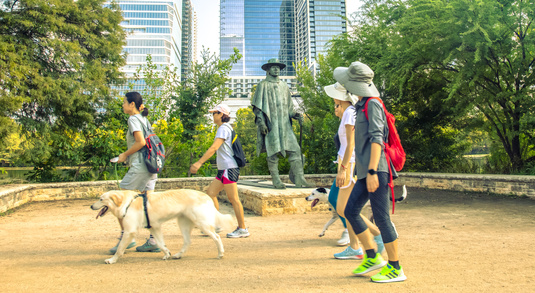 A group of people with dogs walk by the Stevie Ray Vaughan statue along the Ann and Roy Butler Hike-and-Bike Trail near Downtown Austin, Texas