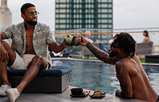Two men, one sitting on a poolside stool and the other in a rooftop pool, clink glasses in a toast at The Otis Hotel in Austin, Texas