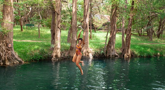 Woman in a bathing suit swinging on a rope swing above the iconic Blue Hole swimming spot in Wimberley, Texas