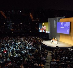 Two people sit in chairs on an auditorium stage before an audience that can also see big screens above the stage in Austin, Texas.