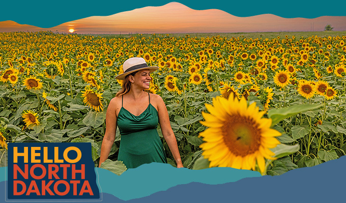 Smiling woman wearing a hat and a green dress walking through a large field of sunflowers with a sunset in the background, with overlay text that reads: Hello North Dakota