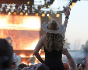 View from behind of a woman wearing a white cowboy hat sitting on someone's shoulders in the crowd of an outdoor concert