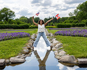 Smiling woman holding US and Canadian flags while standing along a water feature in a garden