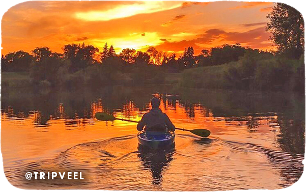 View from behind of a person paddling in a kayak on a body of water with an orange sunset in the background, with overlay text that reads: @tripveel