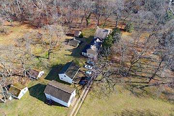 An aerial view of historic home, outbuildings and cars in a wooded clearing.