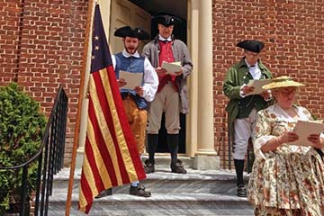 4 costumed historians standing beside an American flag read the Declaration of Independence on exterior steps of building entrance.	