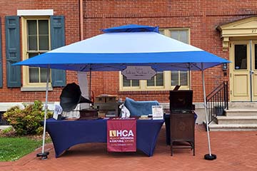 A blue and white covered display table with an HCA logo features victrolas and a statue of the white dog with black ears, Nipper, in front of the Old State House. 	