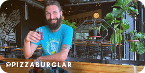 Man holding up a drink inside a restaurant at a wood table, with overlay text that reads: @pizzaburglar