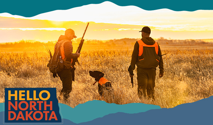 View from behind of two men standing in a field with a dog, holding hunting rifles with sunset in the background, with overlay text that reads: Hello North Dakota
