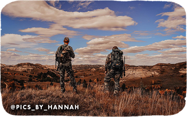 View from behind of two people wearing camo standing in a wide open space looking out to rock formations in the distance, with overlay text that reads @pics_by_hannah