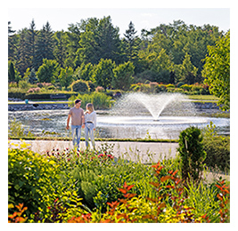 View of a distance of a man and a woman walking among the colorful flowers in front of a fountain at the International Peace Garden