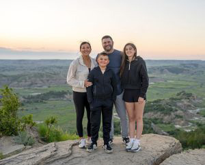 Smiling family of a mom, dad, a young boy and a young girl posing on a cliff with a sunset and scenery of ND in the background