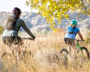 View from behind of two people riding bikes through a meadow with hills in the background