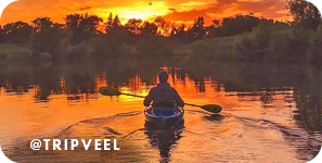 View from behind of a person kayaking on a river with a brilliant orange sunset in the background, with overlay text that reads: @tripveel