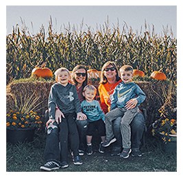 Two women and three young boys posing with hay bales and pumpkins in front of a corn maze