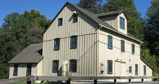 Exterior side view of beige, three-story mill with pitched roof, dormer window and green trim sits at the edge of the road.