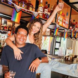 Two people celebrate, raising a glass in a toast, inside a bar adorned with pride flags in Austin, Texas