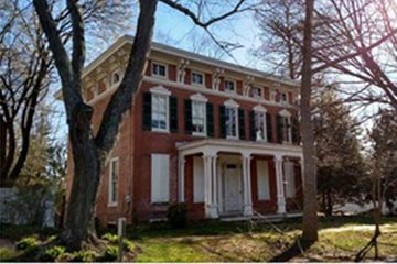 Exterior, side view of three-story brick house with white trim, black shutters and four white columns, surrounded by trees.