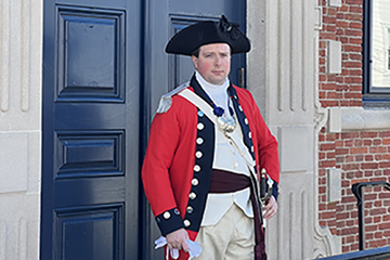 White male wearing traditional British Redcoat attire with black hat and sword on his hip stands in front of brick building with blue door and black iron lighting fixture.	