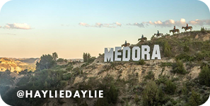 A rocky landscape with horseback riders in the distance and a sign of white letters that reads Medora, with overlay text that reads: @hayliedaylie