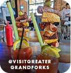 Two bloody marys with long straws and topped with lemons, bacon and other food, with overlay text that reads: @visitgreatergrandforks