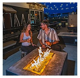 A man and a woman sitting outside around a fire pit at night and drinking craft beers