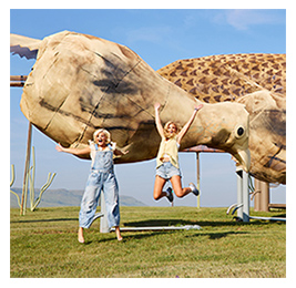 Two women jumping in front of a large sculpture in the middle of a wide-open landscape