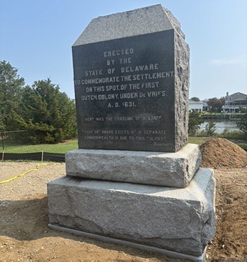 Vertical Monument stands atop two horizontal slabs surrounded by fresh dirt with trees and a body of water in the background.	