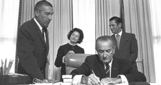 President Lyndon Johnson sits and signs at his desk flanked by two male and one female staffer.	