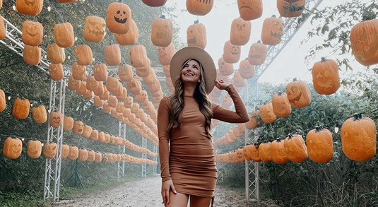  A woman wearing a burnt orange mini dress and a hat strolls through a pumpkin patch aisle underneath hanging jack-o'-lanterns