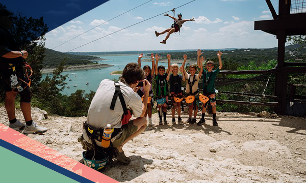 Five children pose for a photo as an adult ziplines in the background at Lake Travis Zipline Adventures near Austin, Texas