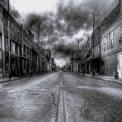 Historic black-and-white photo of an empty street lined with weathered buildings under a dramatic, cloudy sky in Austin, Texas