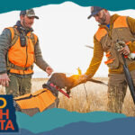 Two men wearing orange and brown hunting outfits with a dog holding a dead bird in a field, with overlay text that reads: Hello North Dakota