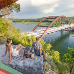 Two people sit on a large rock along a hiking trail at the Pennybacker Bridge Overlook over Lake Austin in Texas