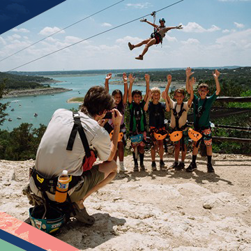 Five children pose for a photo as an adult ziplines in the background at Lake Travis Zipline Adventures near Austin, Texas
