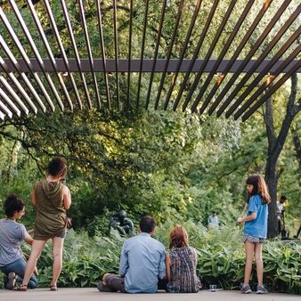 People stand and sit on the ground under a pergola at UMLAUF Sculpture Garden & Museum in Austin, Texas.