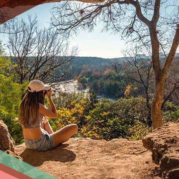A woman aims her camera to take a photo while sitting on a scenic overlooking the Barton Creek Greenbelt in Austin, Texas