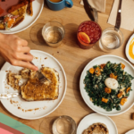 A person's hands use a knife and fork to cut French toast on a table filled with brunch items at Paperboy on South Lamar in Austin, Texas
